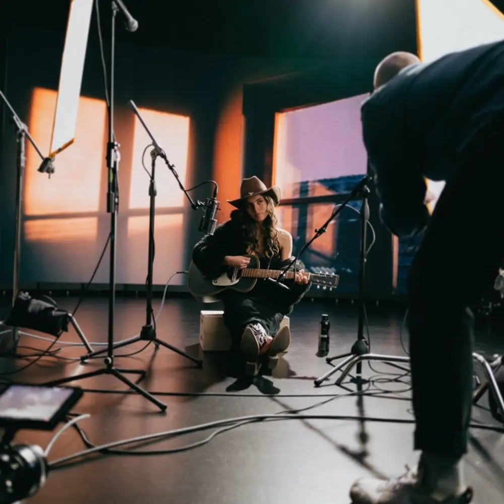 A musician sits on an apple box playing guitar and singing in front of an LED wall while being filmed for a music video
