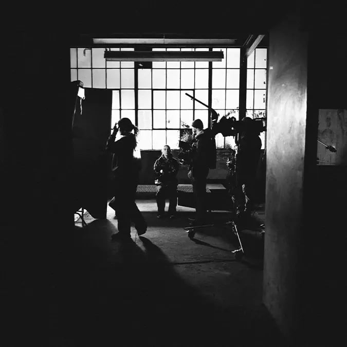 A boxer sits on a bench in an industrial warehouse while a film crew prepares camera and lighting equipment