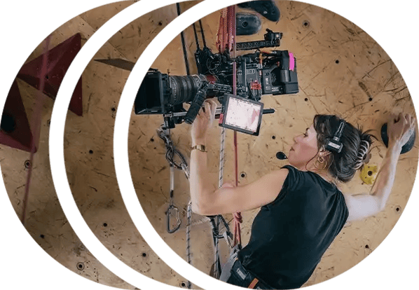 A camera operator operates a RED cinema camera while suspended on a rock wall at a climbing gym